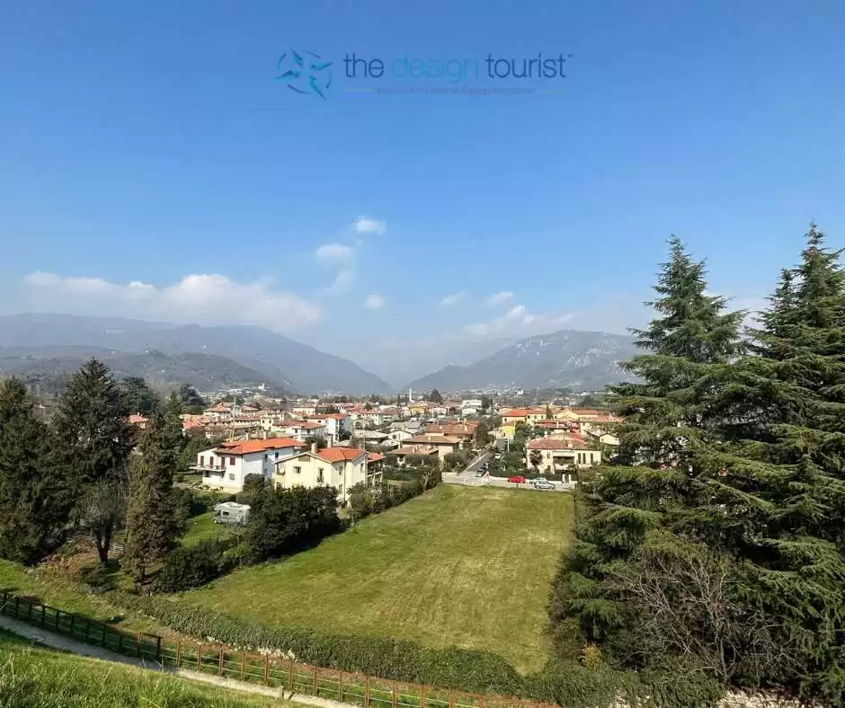 A picturesque mountain town of bassano del grappa with clustered houses on a hillside overlooking a river valley, with mountains in the background under a blue sky.
