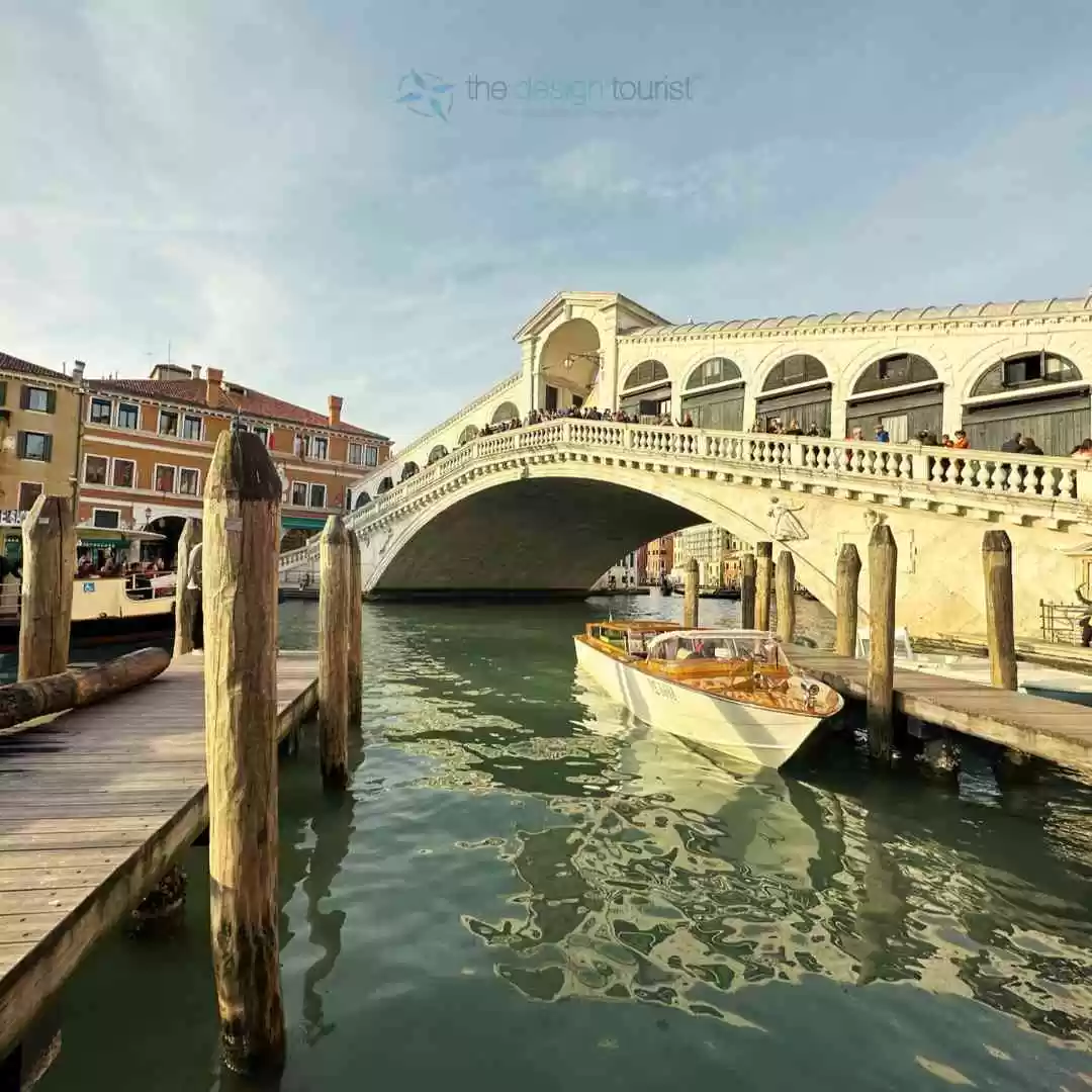 A small boat traveling down a narrow venice canal under Rialto Bridge. The text "The Design Tourist" is superimposed on the image.