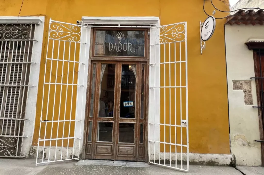 The colorful storefront of Dador, an ethical Cuban fashion boutique in Havana, featuring bright mustard-yellow walls, vintage wooden doors, and open white wrought-iron gates welcoming visitors.