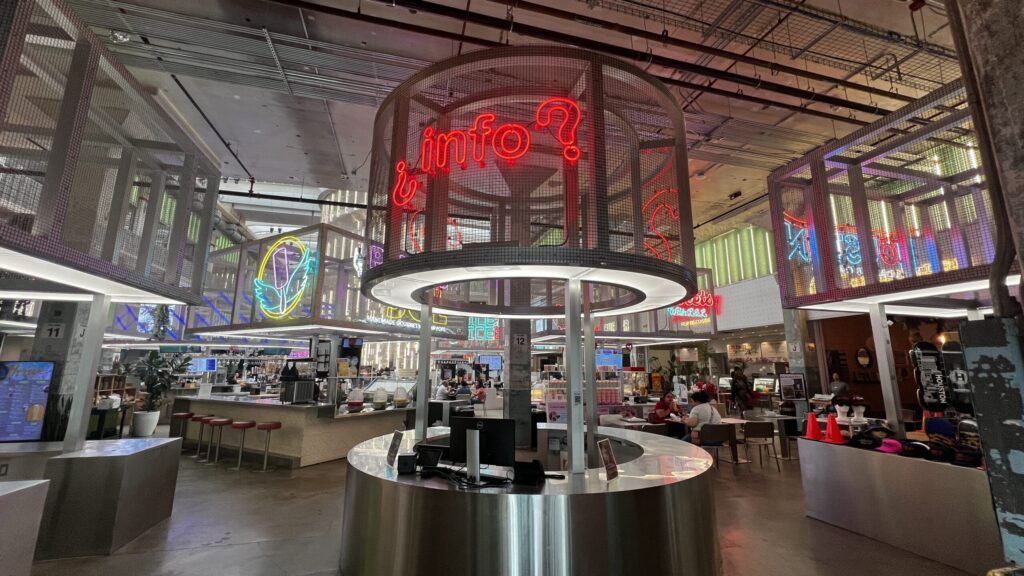 Interior of Post Houston food hall featuring neon signs, modern industrial design, and a central circular info desk surrounded by eateries and seating areas.