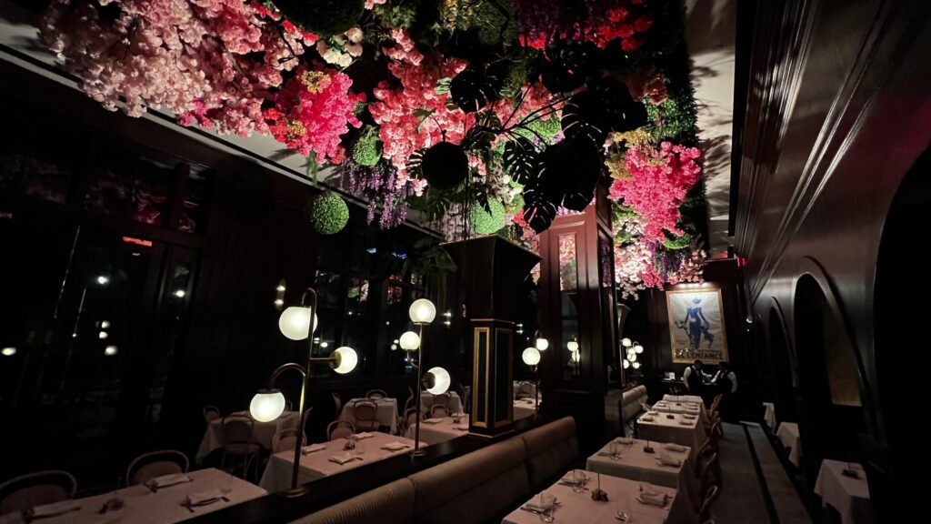 Elegant restaurant interior with white tablecloth dining tables, globe pendant lamps, and a dramatic ceiling installation of lush greenery and cascading pink, red, and purple flowers - experiencing culinary culture in Bayou City.