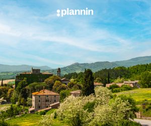 Scenic countryside village surrounded by green hills, trees, and historic stone buildings under a bright blue sky with the text “plannin” above.