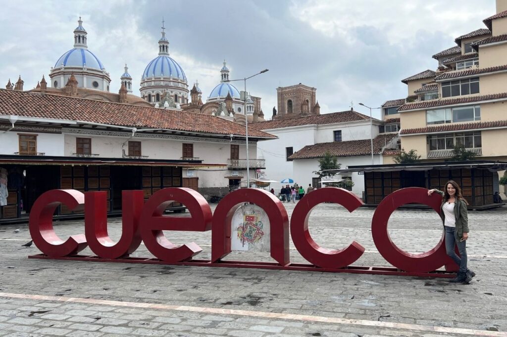 Karen LeBlanc poses next to a large red "Cuenca" sign in a cobblestone city plaza with a historic blue-domed cathedral behind her.