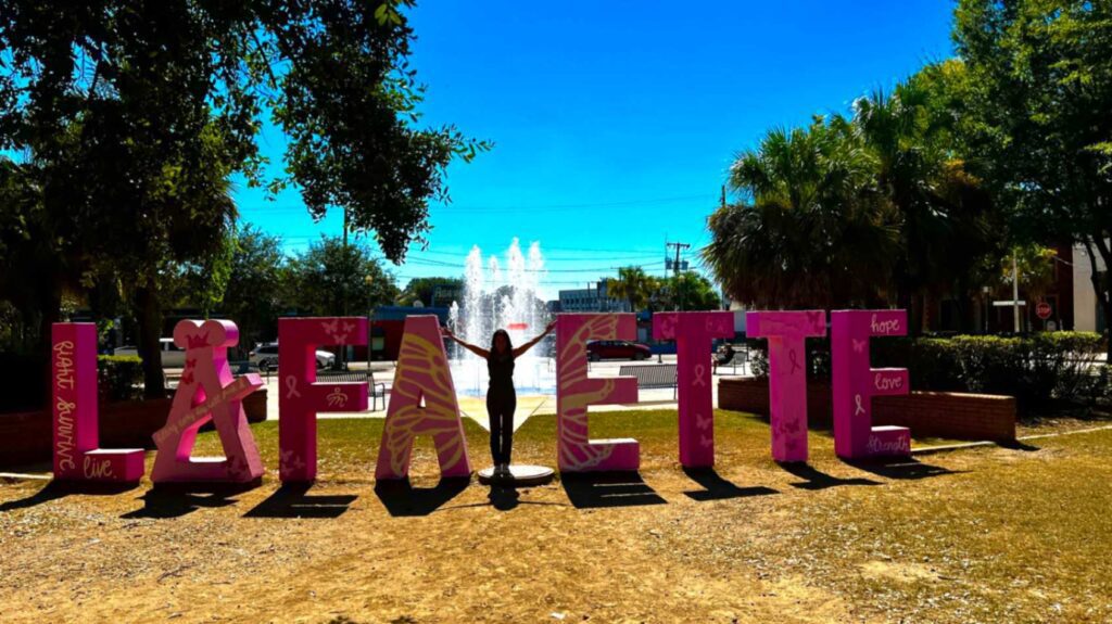 A person poses with arms raised to act as the letter "Y" inside the giant pink breast cancer awareness "LAFA_ETTE" sign in front of a water fountain at Parc Sans Souci.