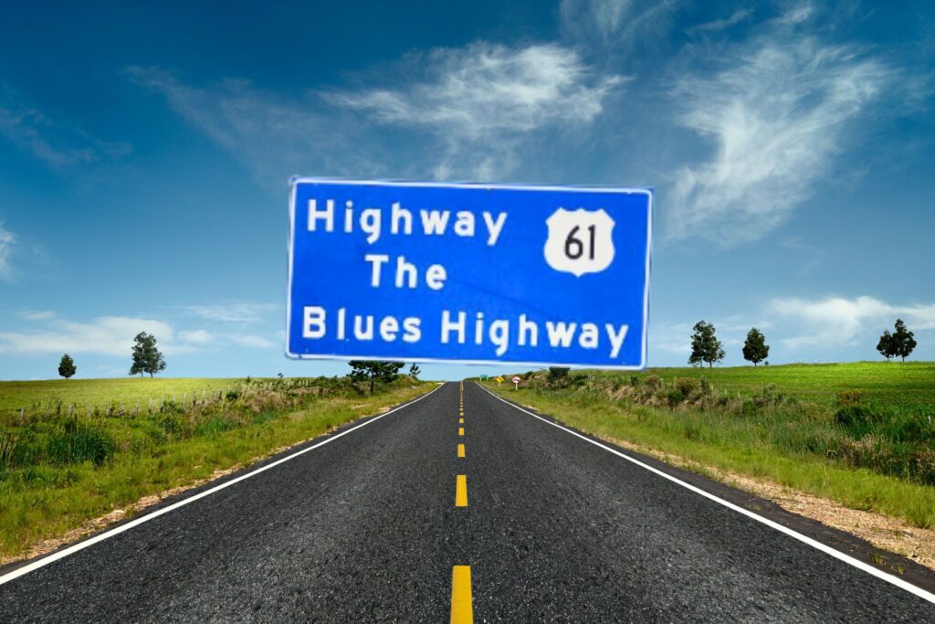 An image of a large, rectangular blue roadside sign with white text that reads "Highway 61 / The / Blues Highway". The number "61" is enclosed inside a white shield icon, representing a U.S. Route shield. The sign is supported by three metal posts and stands along the side of a paved highway. In the background, there is a grassy slope, a line of green trees, and a clear, light blue sky.