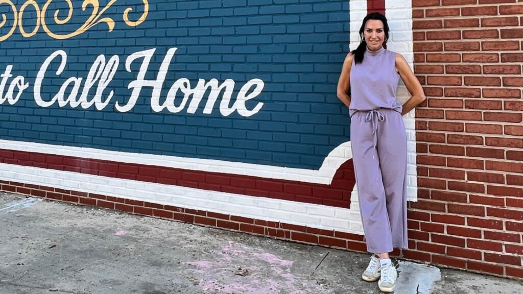 A woman stands smiling in front of a blue and red brick wall painted with white cursive lettering that reads "to Call Home." She is wearing a matching sleeveless lavender or light purple top and drawstring pants outfit with white sneakers.