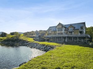 Large three-story wooden lodge built along a green grassy shoreline next to calm water under a blue sky.