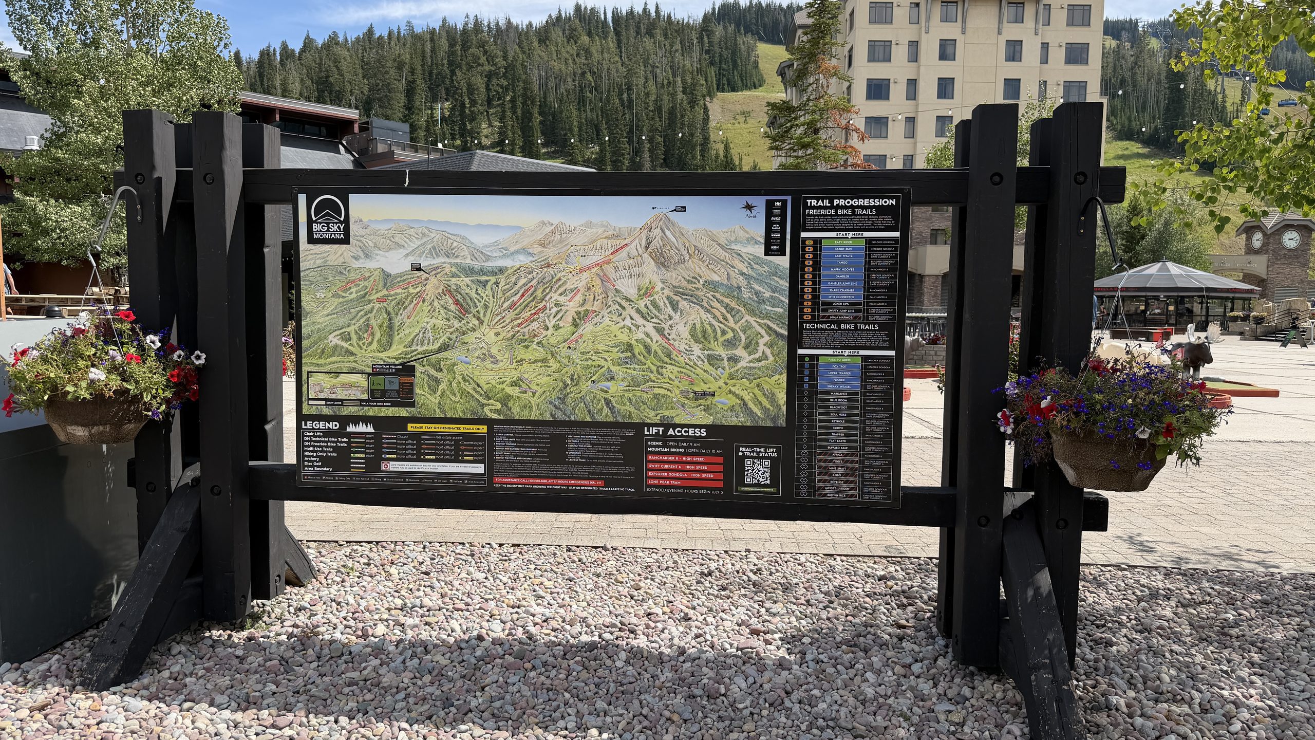 Big Sky Resort summer mountain bike trail map sign on a black wooden frame with hanging flower baskets at the base village in Montana.