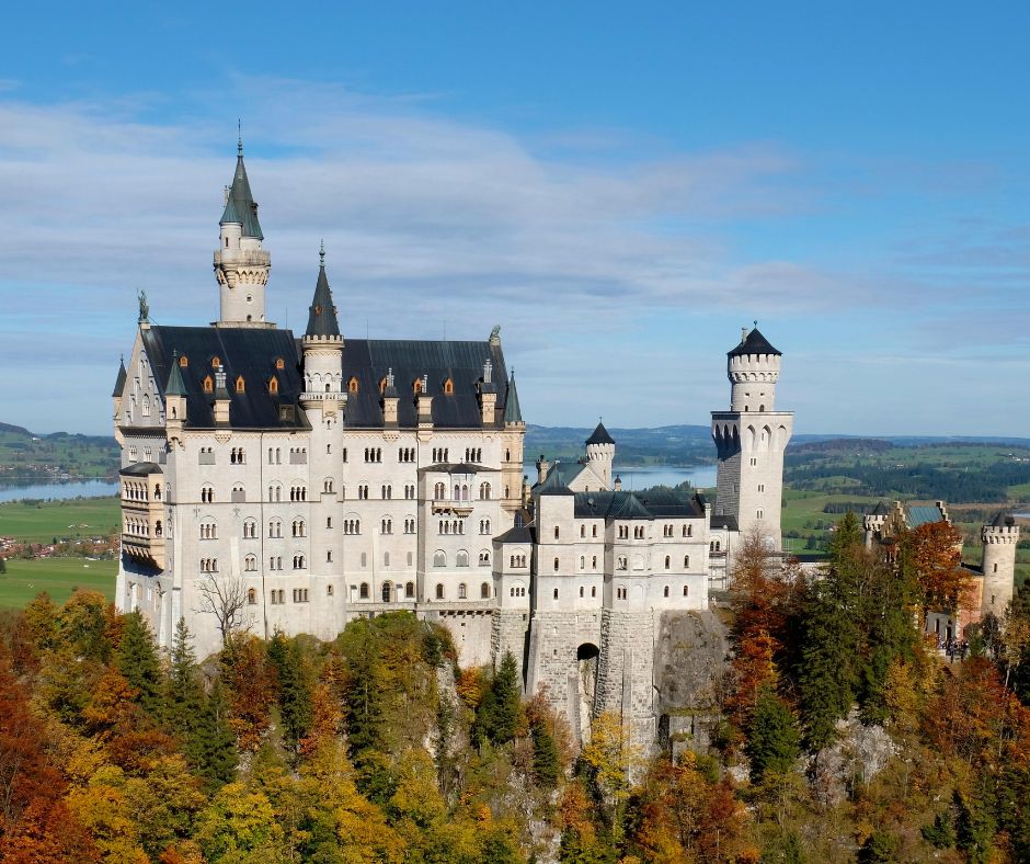 Neuschwanstein Castle standing on a rugged hill surrounded by autumn trees and rolling green hills under a partly cloudy sky
