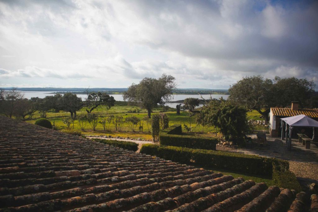 View from a terracotta tiled roof overlooking green fields, olive trees, and Alqueva Lake in Alentejo Portugal under a cloudy sky.